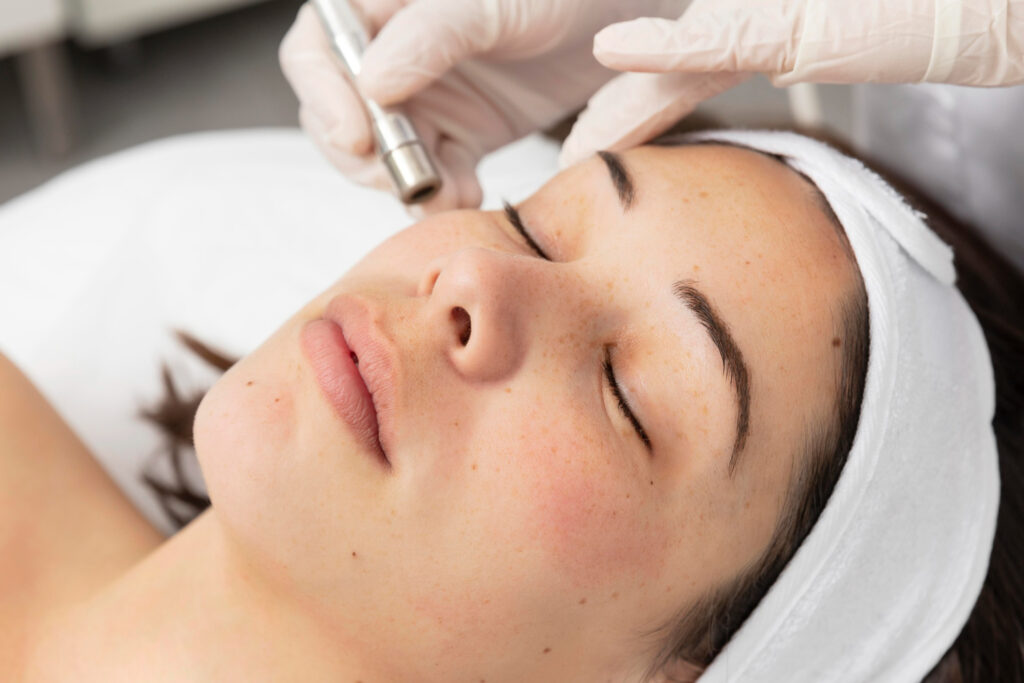 A person is receiving a facial treatment with a skincare device, lying relaxed with closed eyes and a headband, in a clinical or spa setting.