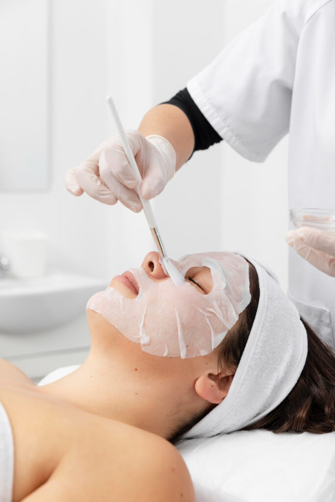 A person in a spa setting is receiving a facial treatment; another person applies a mask with a brush, both in a clean, white environment.