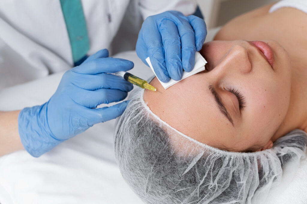 A person in a medical cap is performing a procedure on the eyebrow of a relaxed person lying down, both wearing protective gear.