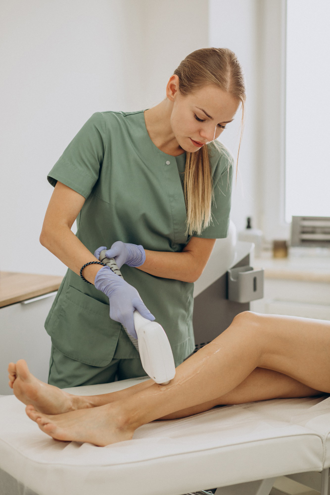 A focused person in a green scrub is performing a procedure using a white device on another person's leg in a clinical environment.