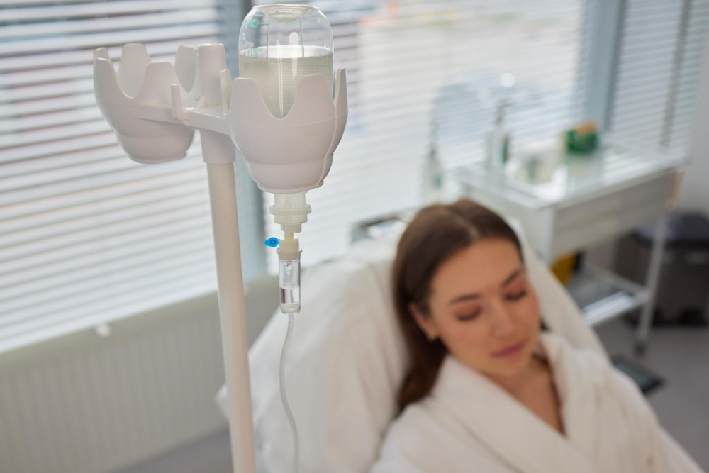 An IV drip stand in focus with a saline bag, leading to a blurred background where a person appears to be resting in a medical setting.