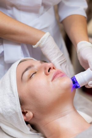 A person in a head wrap reclines while another person dressed in white performs a facial treatment using a device emitting blue light.