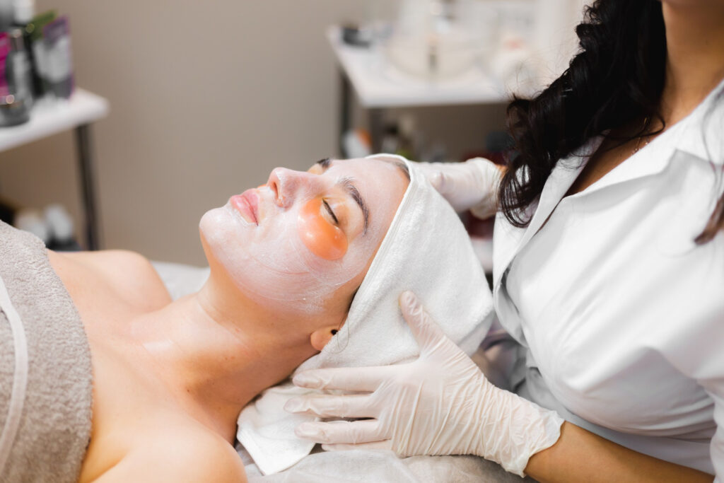 A person is receiving a facial treatment, wearing eye pads, with a skincare professional attending to them in a well-lit spa setting.