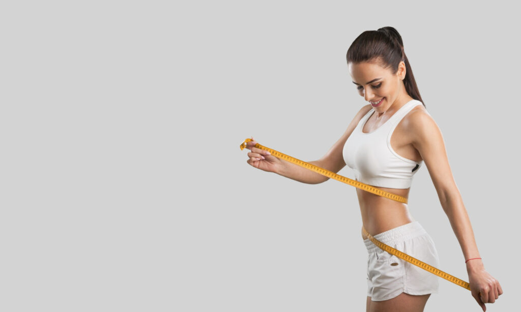 A person is measuring their waist with a yellow tape measure against a gray background, suggesting a concept related to fitness or weight management.