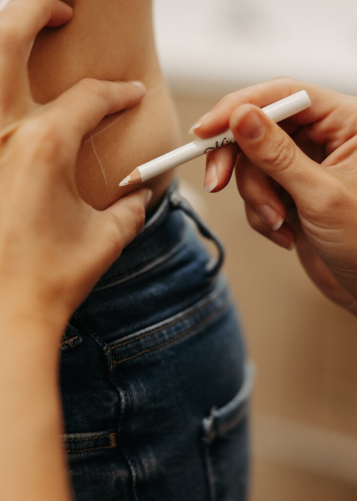 A person marks another's lower back with a white pencil for measurement purposes, wearing denim jeans, in a close-up shot.