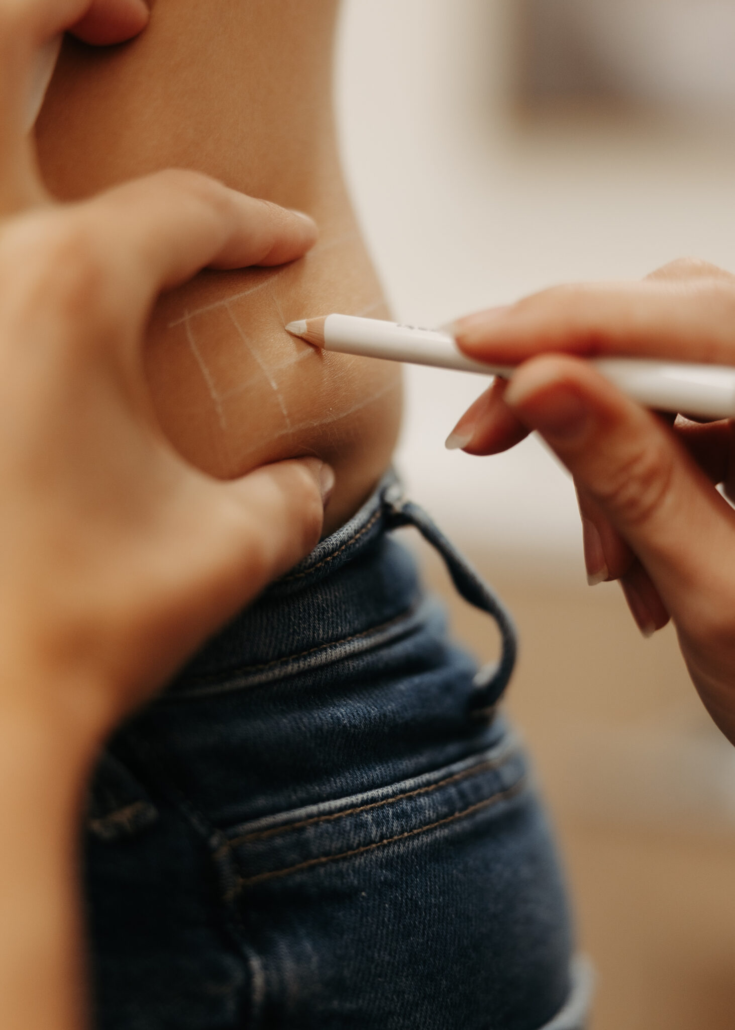 A person draws white lines on skin using a pencil. The focus is on the lower back with denim jeans visible.
