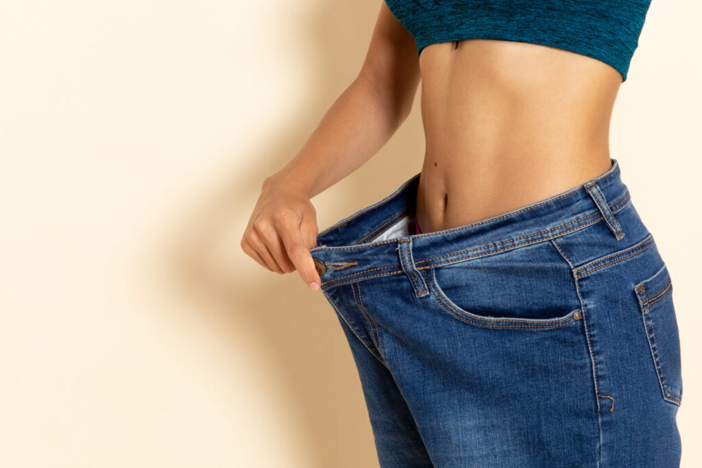 A person is holding out oversized blue jeans, showing a significant gap at the waist, implying weight loss, against a beige background.