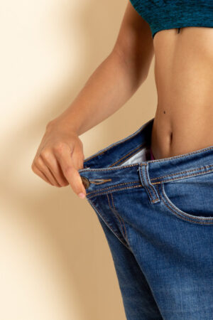 A person is holding out oversized blue jeans, showing a significant gap at the waist, implying weight loss, against a beige background.