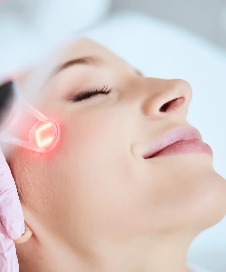 A person is receiving a skincare treatment with a red light device, targeting the cheek area. The setting appears clinical or spa-like, with a focus on rejuvenation.
