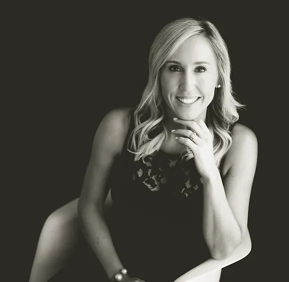 A person with blonde hair and a bright smile sits against a dark background in a monochrome photo, resting their chin on their hand.