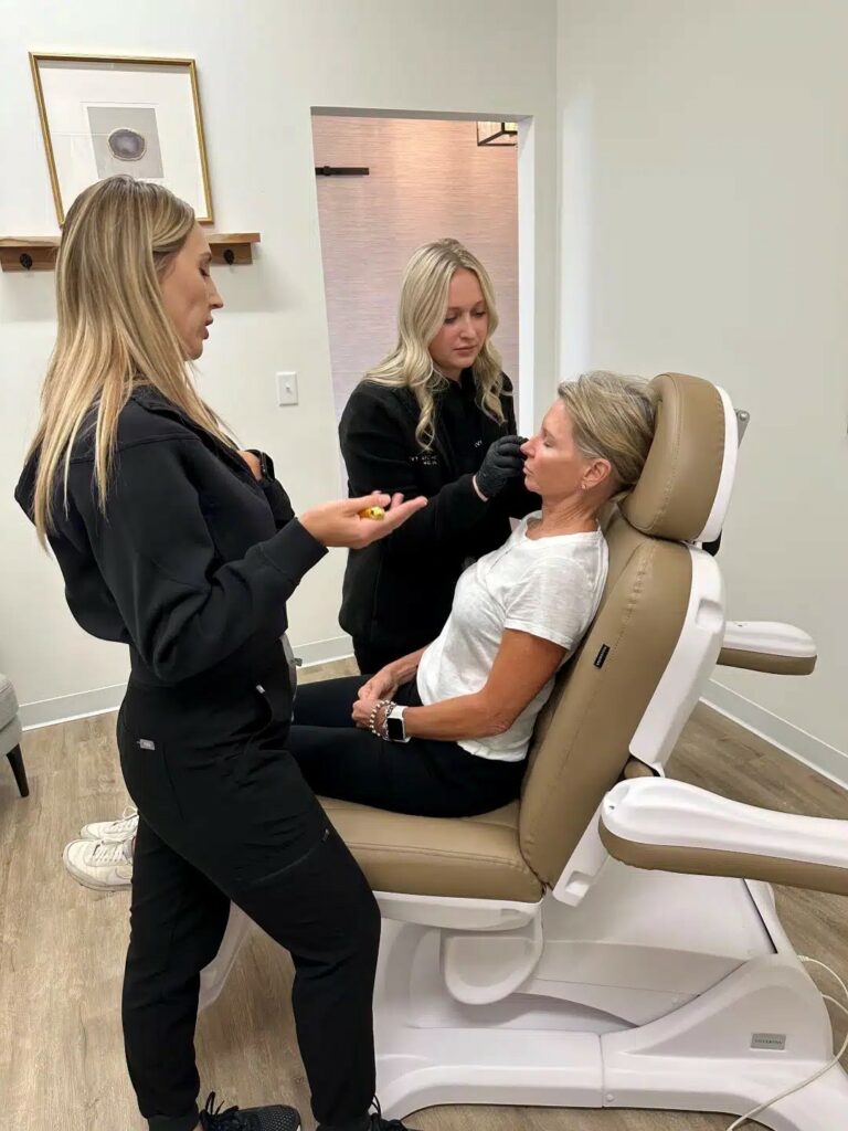 In a clinical setting, two standing persons in black attire appear to be consulting with a seated person, possibly during a medical or cosmetic appointment.