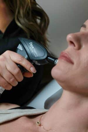 In the image, a person is performing a skin treatment on another person's face using a handheld medical device. The recipient appears relaxed.