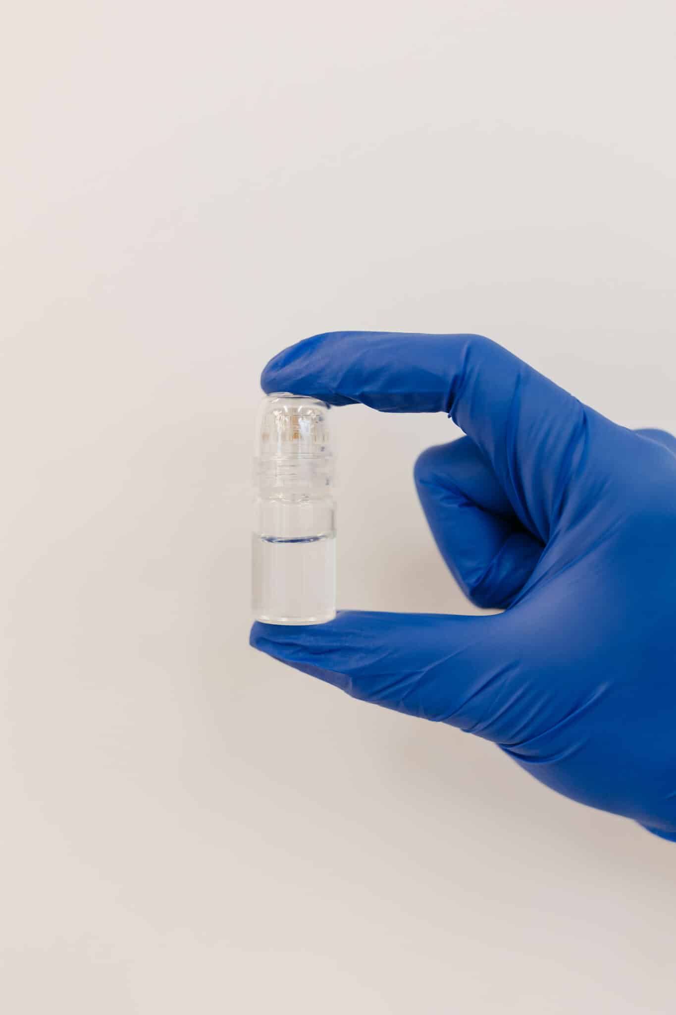 A person wearing a blue glove holds a small transparent bottle with a dispenser, against a plain white background.