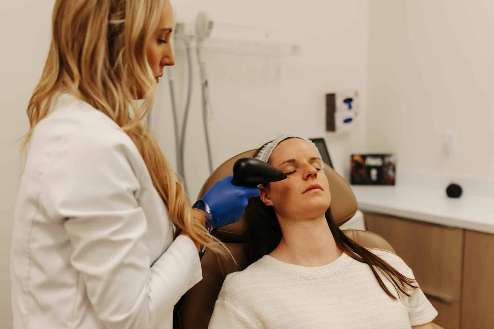 A person in a white coat uses a handheld device on another person's face, who is reclining with eyes closed, possibly receiving a skincare treatment.