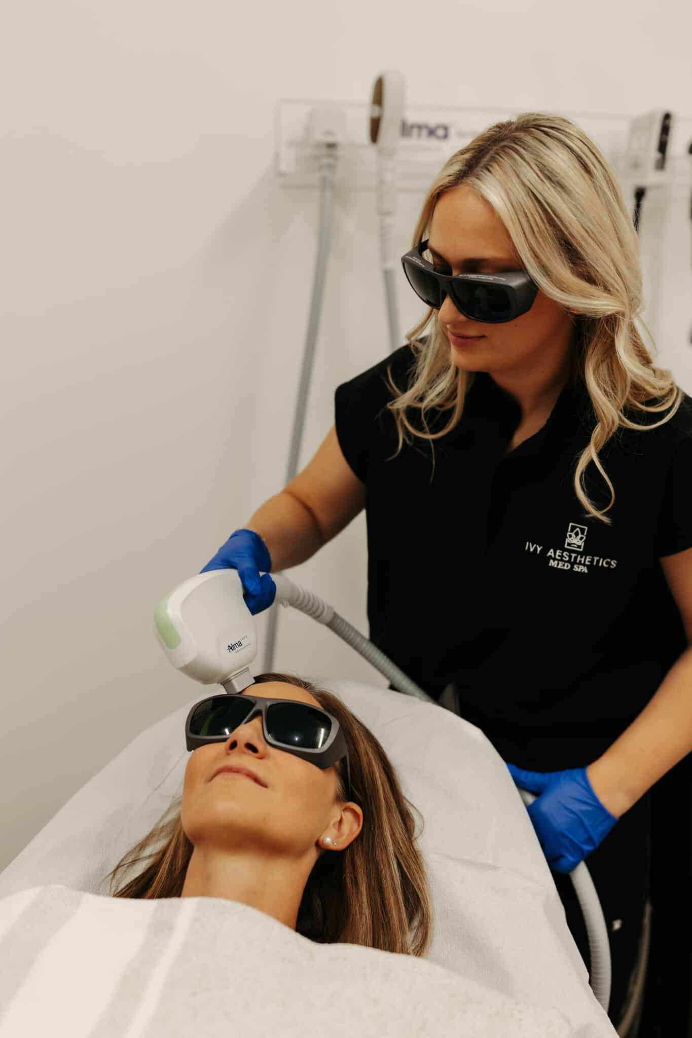 A person is performing a laser skin treatment on a reclining client wearing protective eyewear, in a clean, clinical setting with professional equipment.