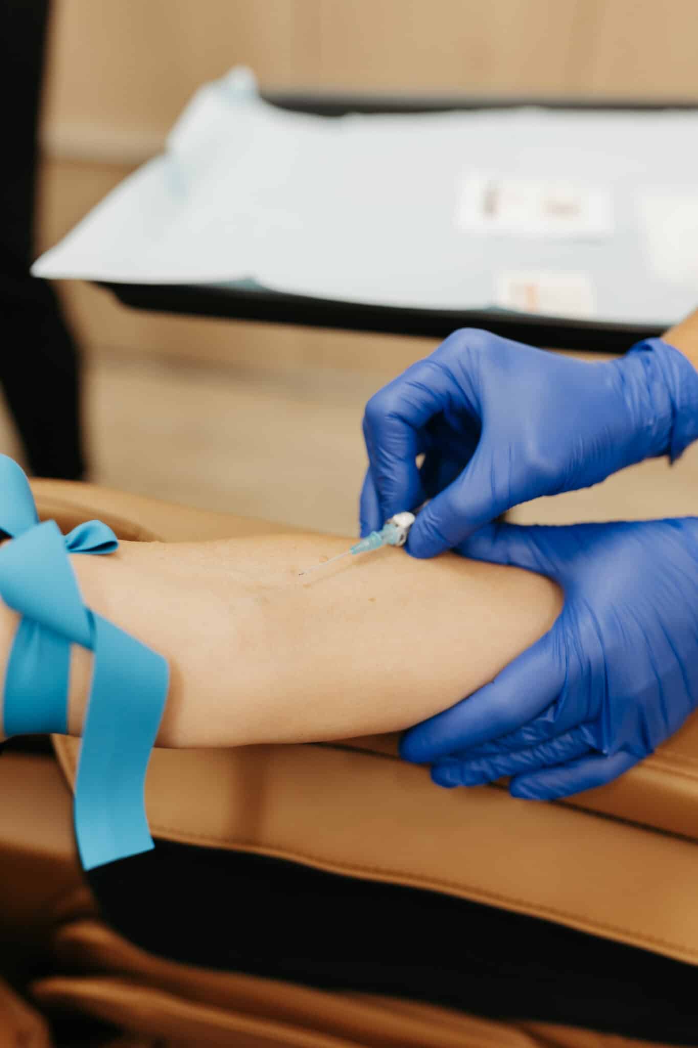 A healthcare worker wearing blue gloves administers an injection into a person's arm, which is tied with a blue tourniquet.