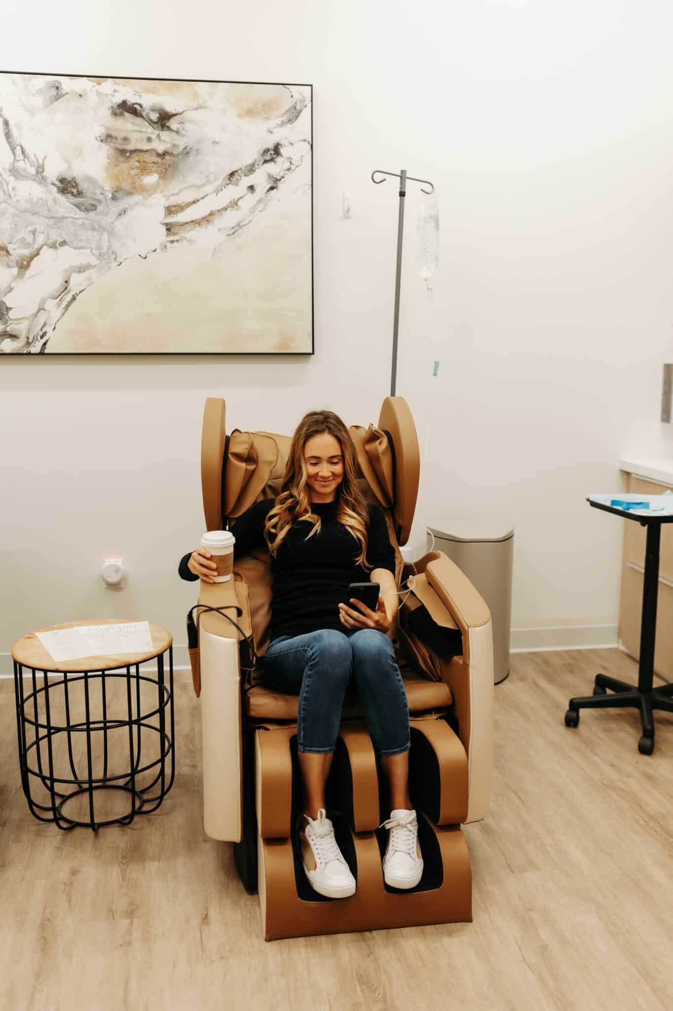 A person is sitting in a medical recliner, looking at a phone, with an IV stand nearby, in a room with modern decor and abstract art.