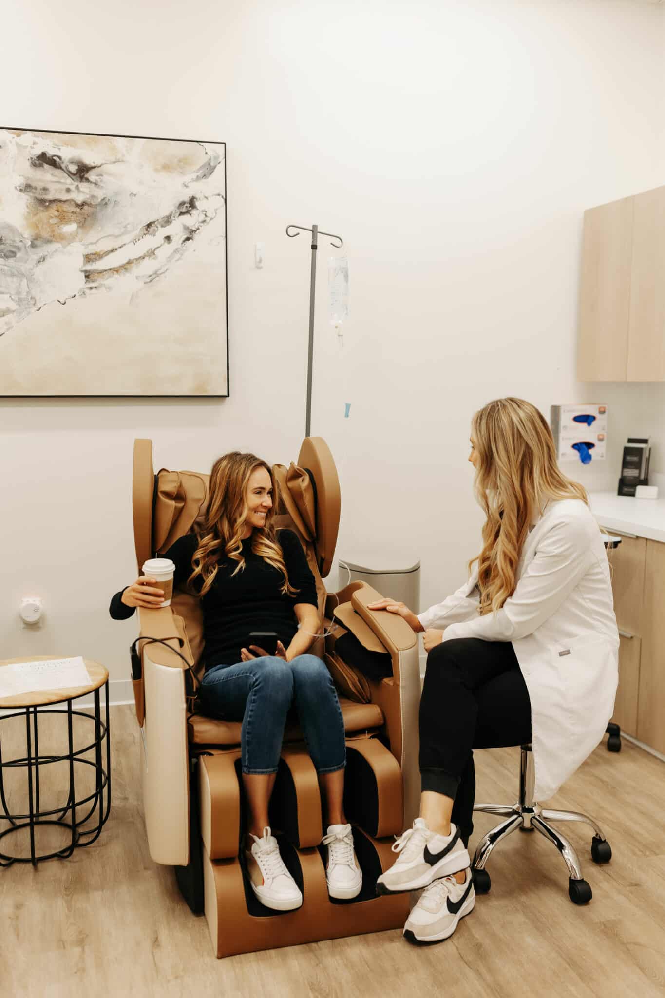 A person in a black top sits in a brown medical chair, holding a cup and phone, conversing with a healthcare professional in a white coat.