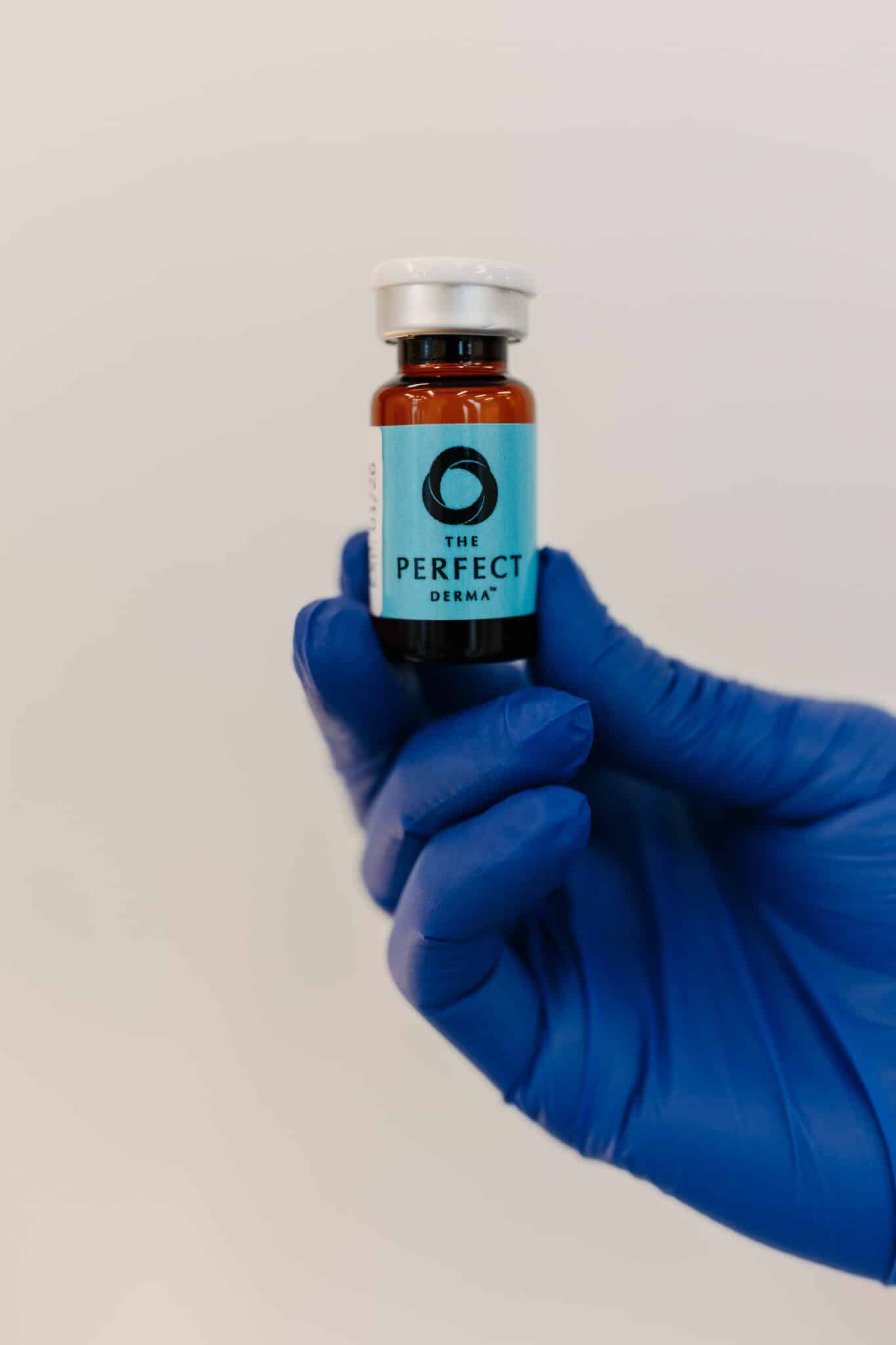 A person wearing blue gloves holds a small amber bottle labeled "THE PERFECT DERMA" against a pale background. The bottle has a white cap.