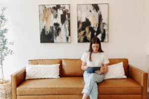A person is seated on a brown leather couch, focused on a tablet, with abstract art and a plant in the background, indoors in a well-lit room.