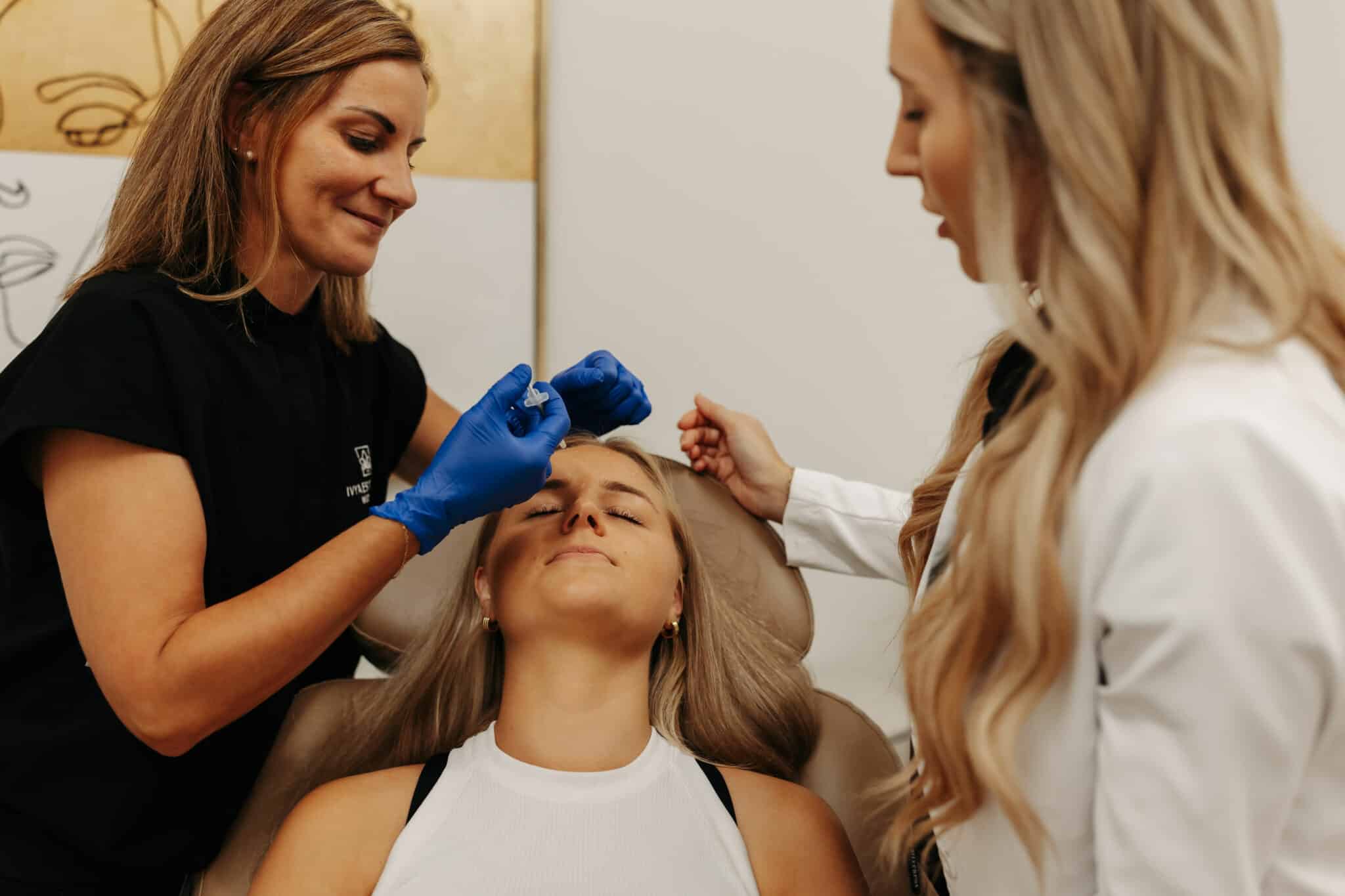 In a well-lit room, a person is performing a facial treatment on a reclining client, with another person assisting or observing. Everyone appears focused and professional.