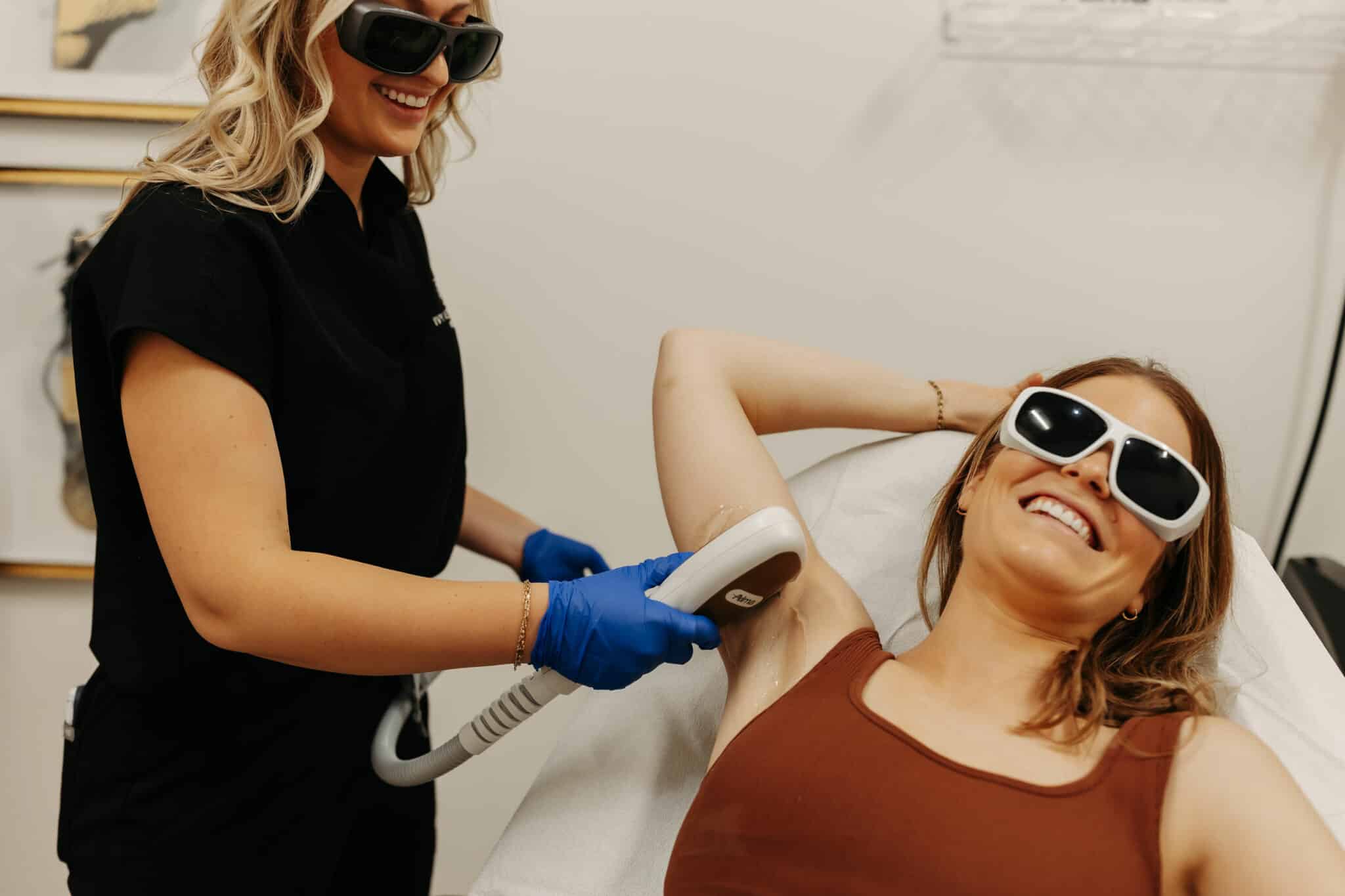 A person is performing a laser treatment on another individual's underarm. Both are wearing protective eyewear, and the client appears relaxed and smiling.