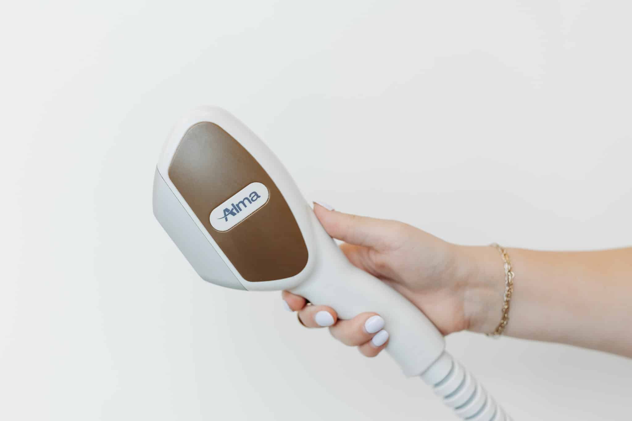 A person's hand is holding a professional hair removal laser device with a brown treatment head against a white background, indicating cosmetic skin treatment.