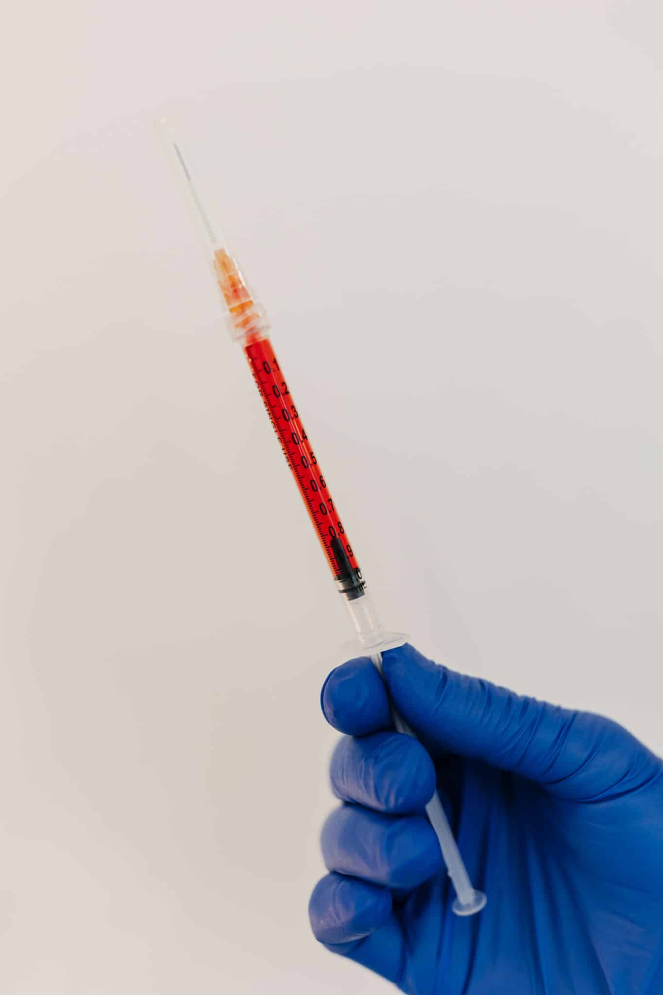 A person's gloved hand holds a syringe filled with a red substance against a white background, suggesting a medical or laboratory setting.