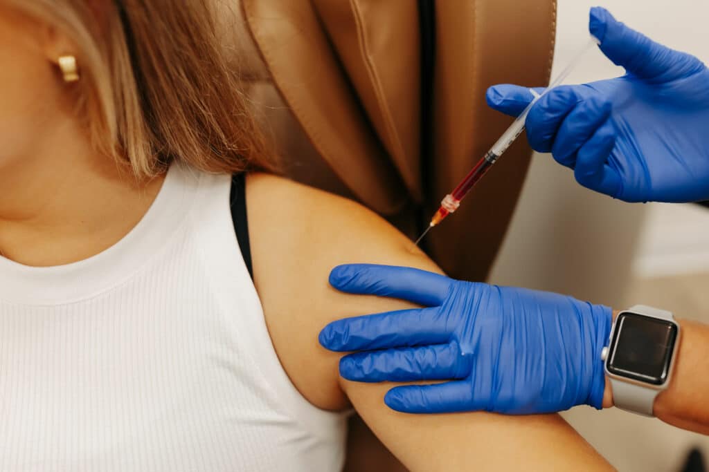 A healthcare professional wearing blue gloves administers a vaccine to a person's upper arm. The person wears a white sleeveless top and a smartwatch.