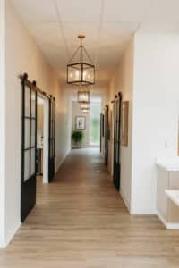 A modern hallway with wooden floors, framed glass doors, elegant pendant lights, wall art and a plant at the end, with a minimalist design.