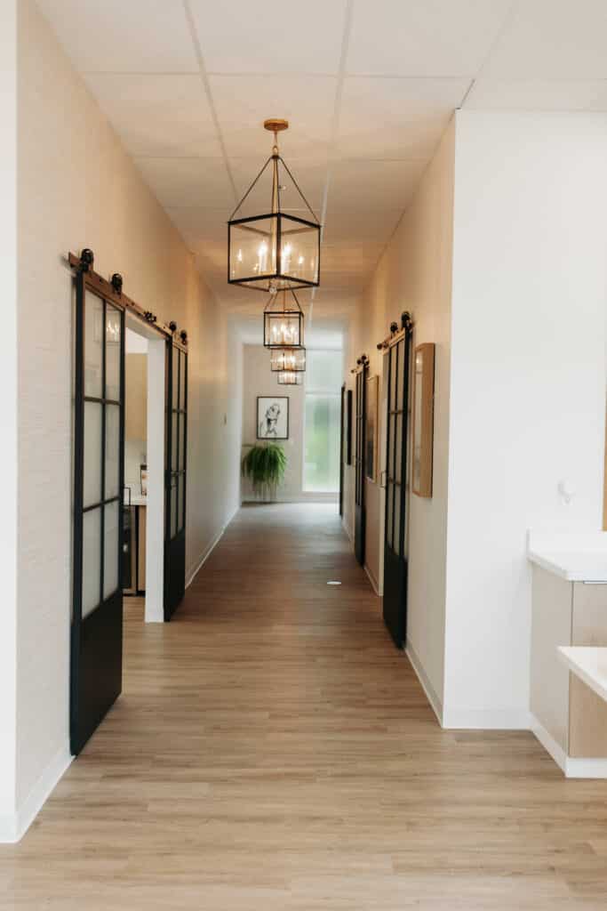 A modern hallway with wooden floors, framed glass doors, elegant pendant lights, wall art and a plant at the end, with a minimalist design.