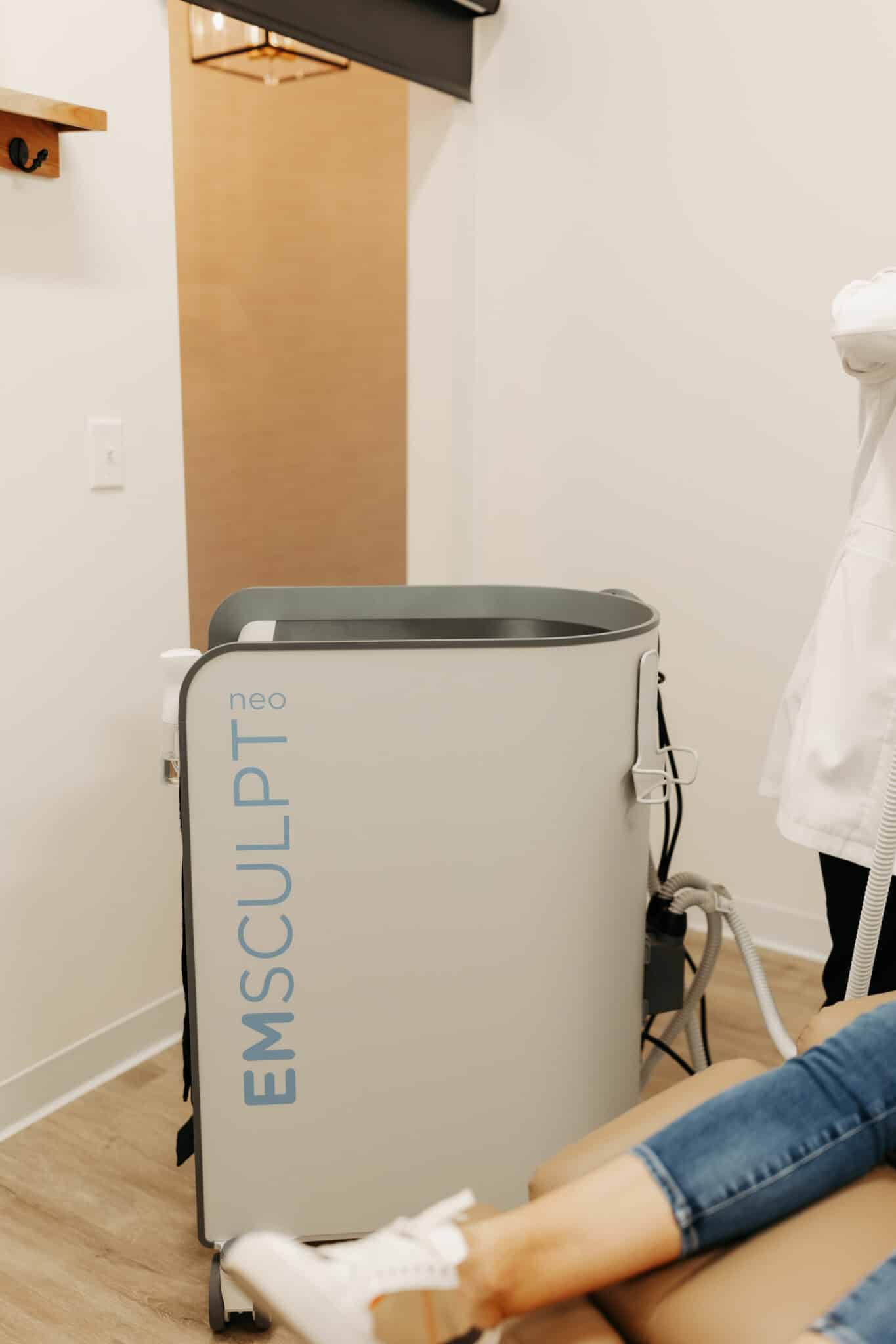 A medical device with "EMSCULPT" branding is featured next to a person in a clinical setting. The person is partially visible, wearing denim and sneakers.