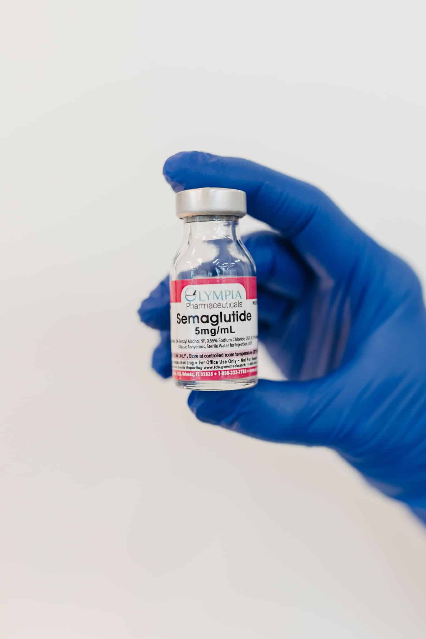 A hand wearing a blue glove holds a vial labeled "Semaglutide 5mg/mL" against a white background. The vial appears to be a medication.