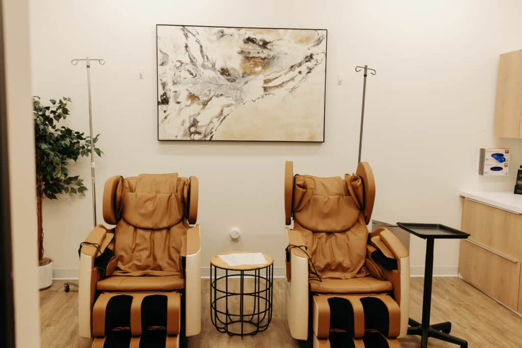 The image shows a modern, clean medical or spa room with two brown massage chairs, an abstract painting on the wall, and IV stands.
