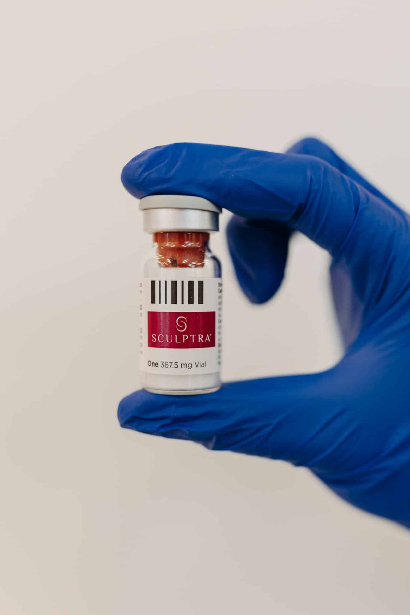 A person wearing a blue glove holds a SCULPTRA vial against a white background. The vial is labeled as containing 367.5 mg.