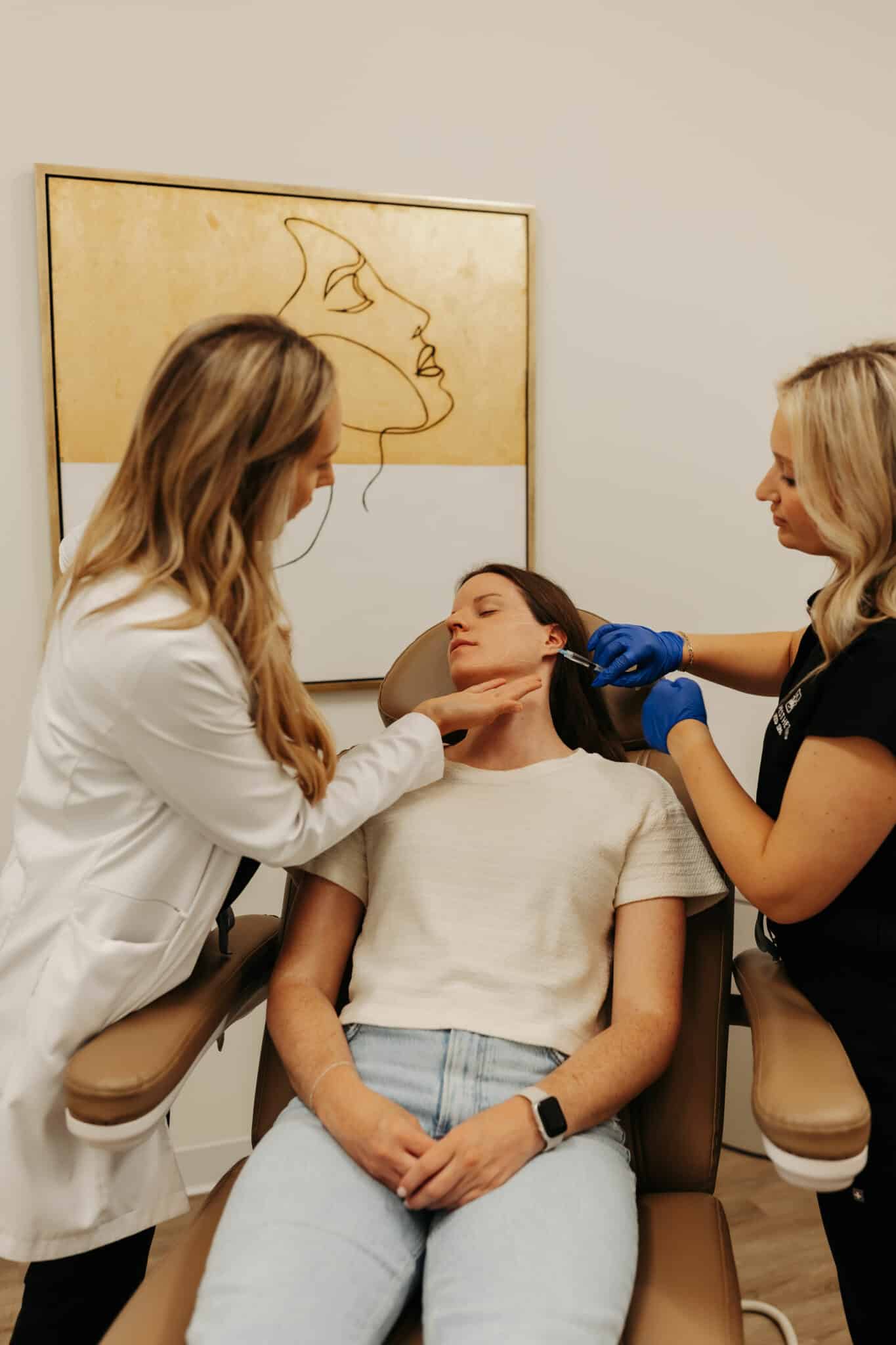 Two persons in medical attire are performing a procedure on a seated person's face in a clinical setting with a minimalist art piece in the background.