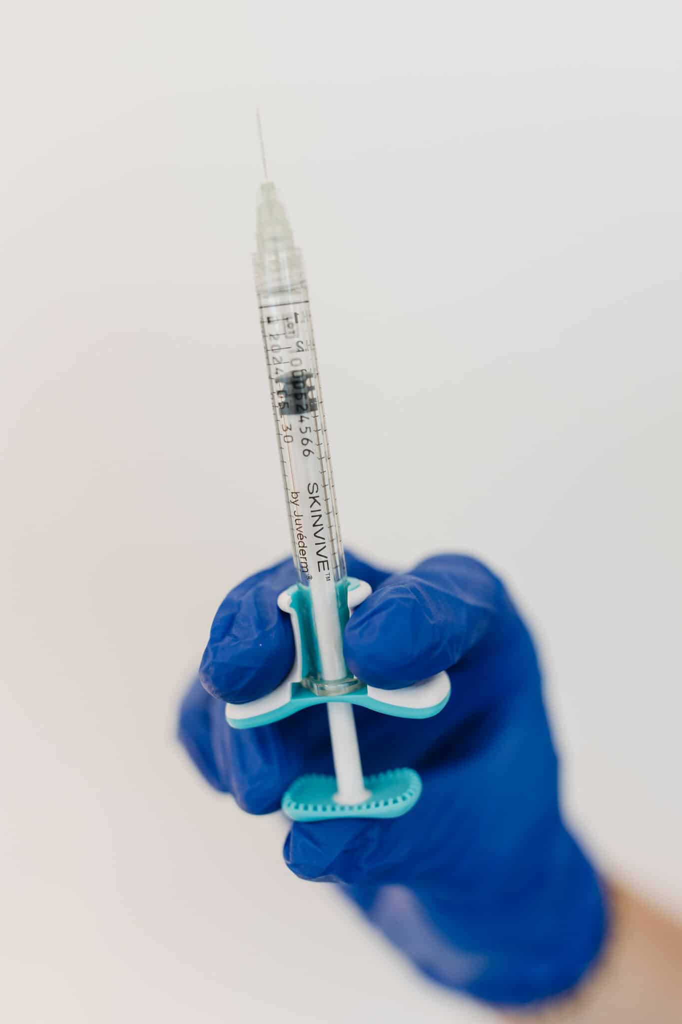 A person's hand in a blue glove holds a syringe against a light background, indicating a medical or healthcare setting, likely for injection.