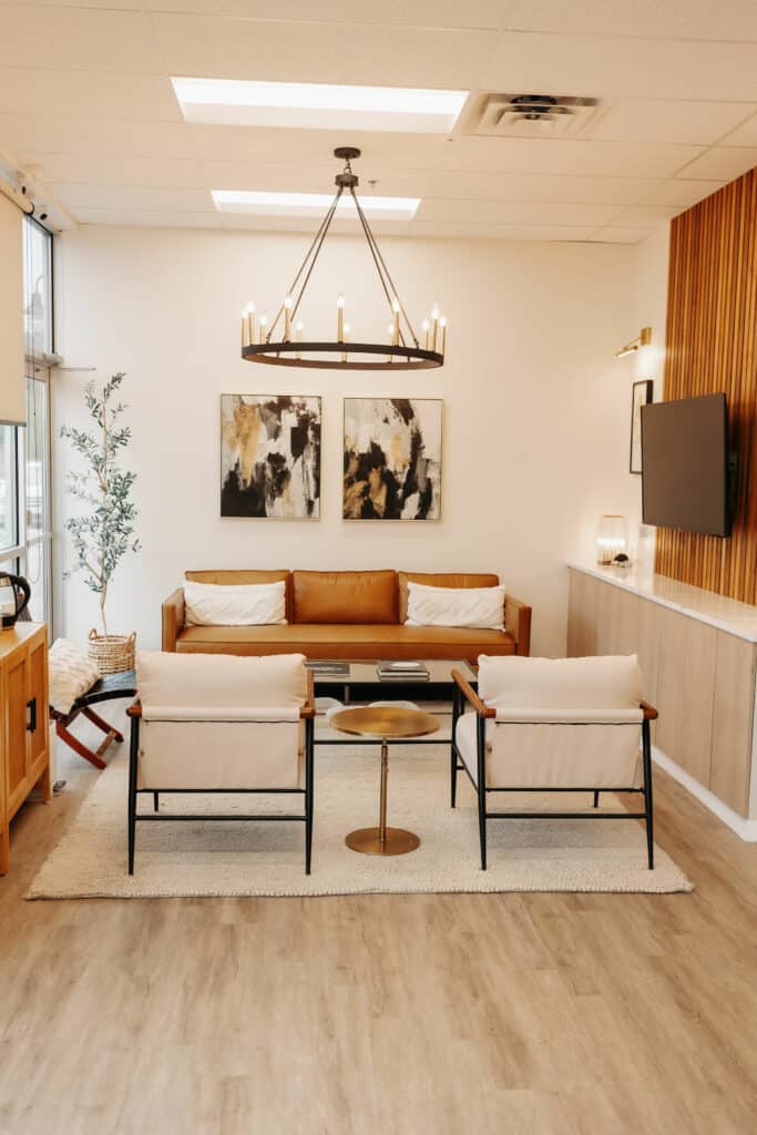 Modern living room with tan leather couch, two armchairs, round coffee table, abstract wall art, chandelier, wooden sidings, and potted plant.