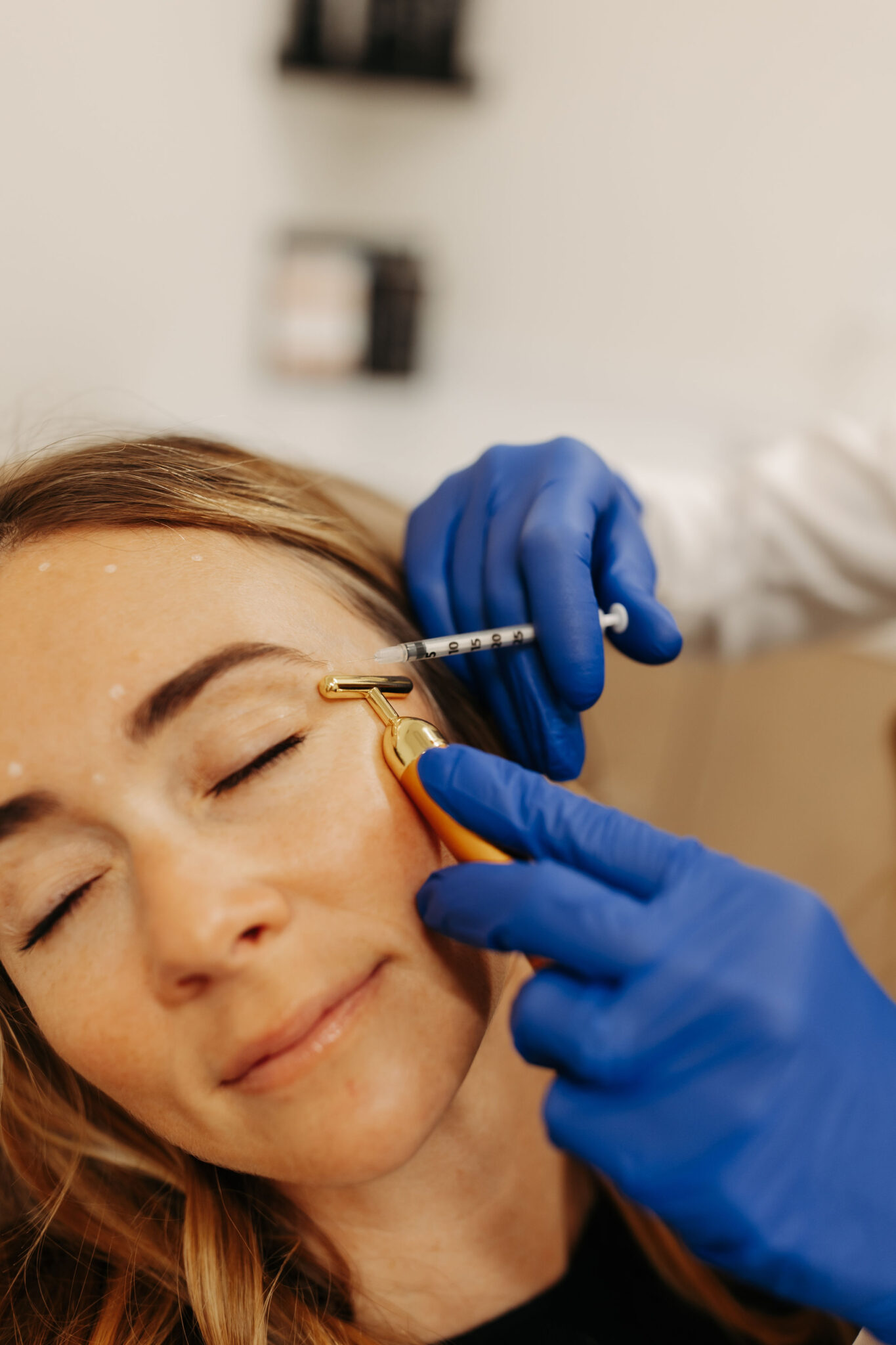 A person receives a cosmetic treatment with blue-gloved hands holding a syringe and a gold-toned facial tool.