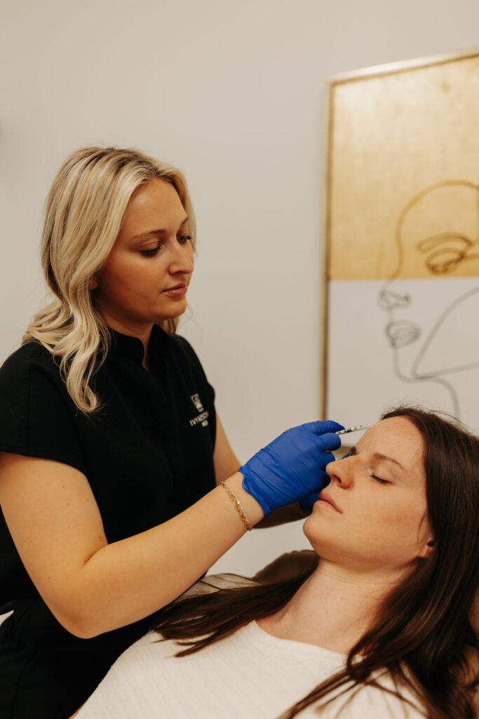 A person in blue gloves cosmetically injects another person's forehead in a clinic setting, with abstract art in the background.