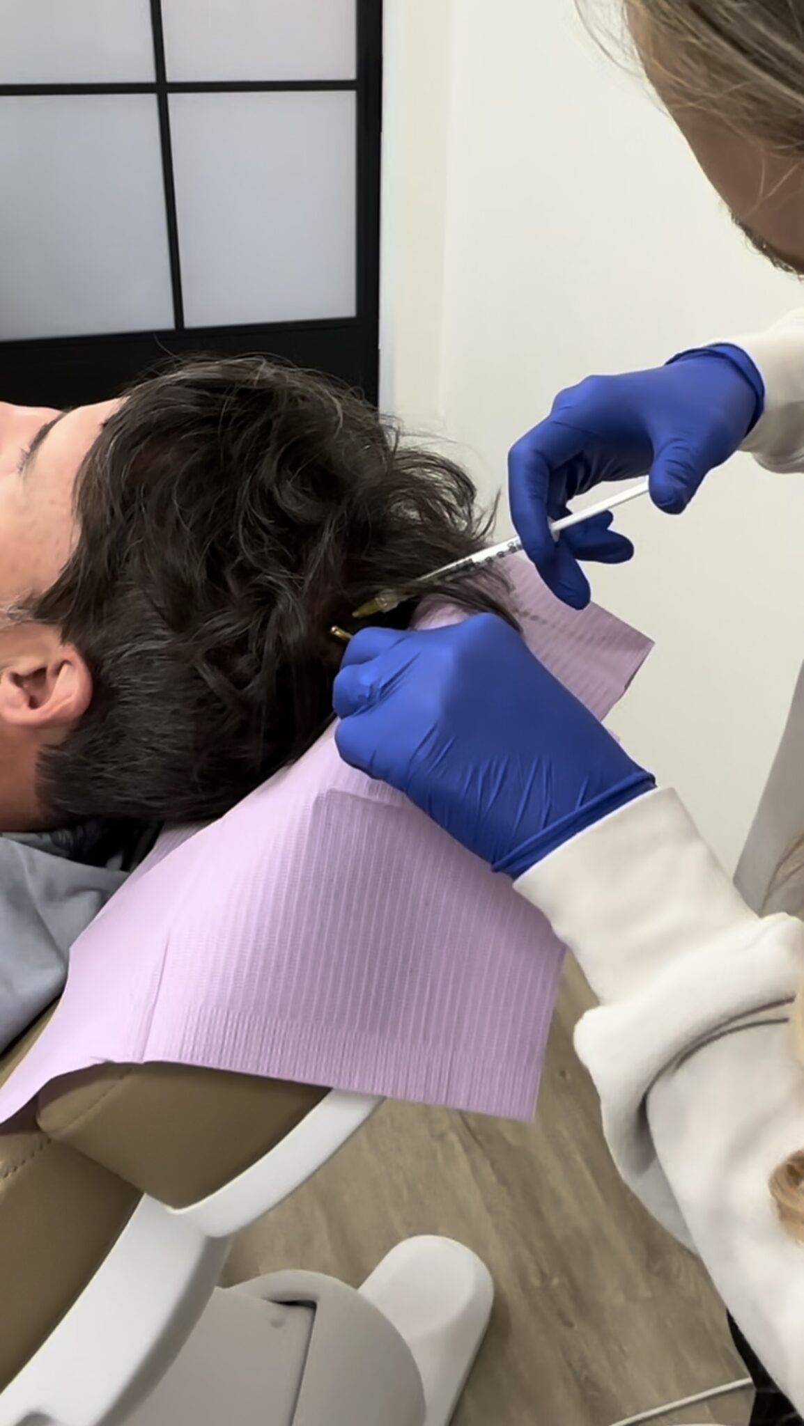 A person in a reclining chair receives a scalp treatment from another person wearing blue gloves. A purple neck bib is visible.