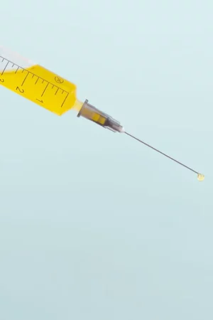An isolated syringe with a yellow plunger and clear barrel containing a liquid is depicted against a plain light background, with a needle extending outwards.