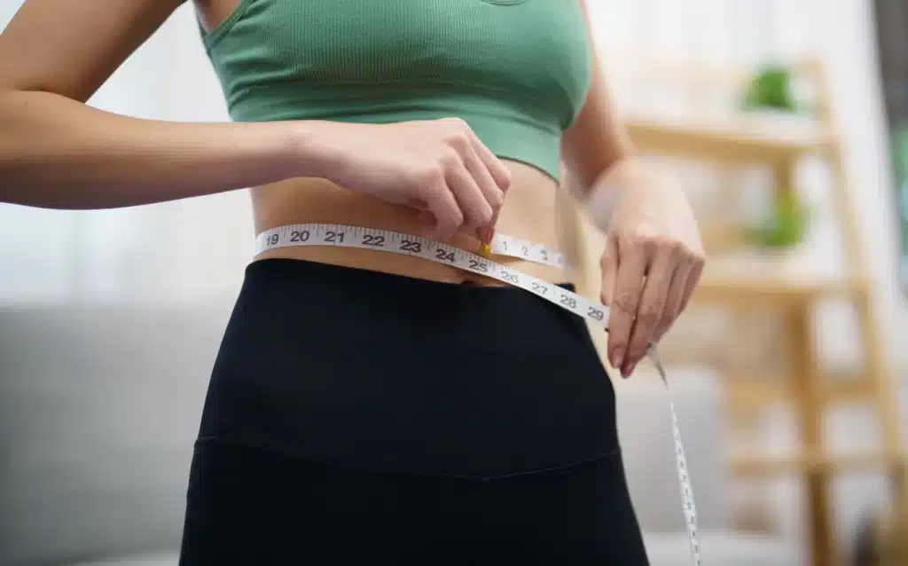 A person is measuring their waist with a white measuring tape against a blurred background, suggesting a theme of health, fitness, or weight management.
