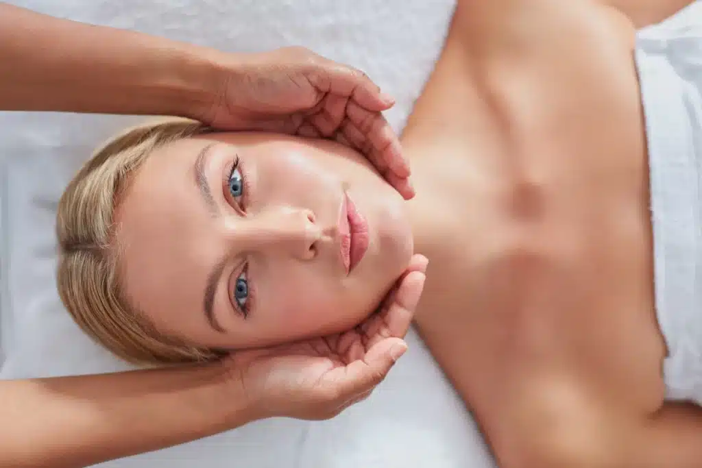 A person is receiving a facial massage, lying down with eyes open, looking relaxed. Two hands gently hold the person's face, suggesting a spa treatment.