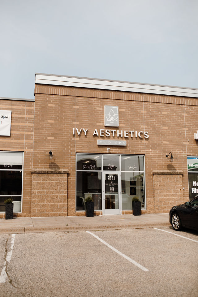 A brick storefront with signage reading "IVY AESTHETICS MED SPA" and "2681" over a glass door, flanked by black potted plants and parked cars.
