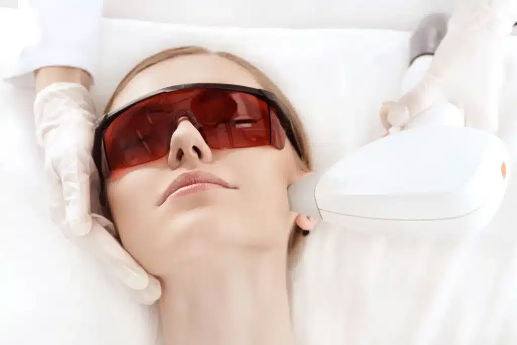 A person lies with red protective glasses while a gloved hand operates a laser device near their face in a clinical setting.