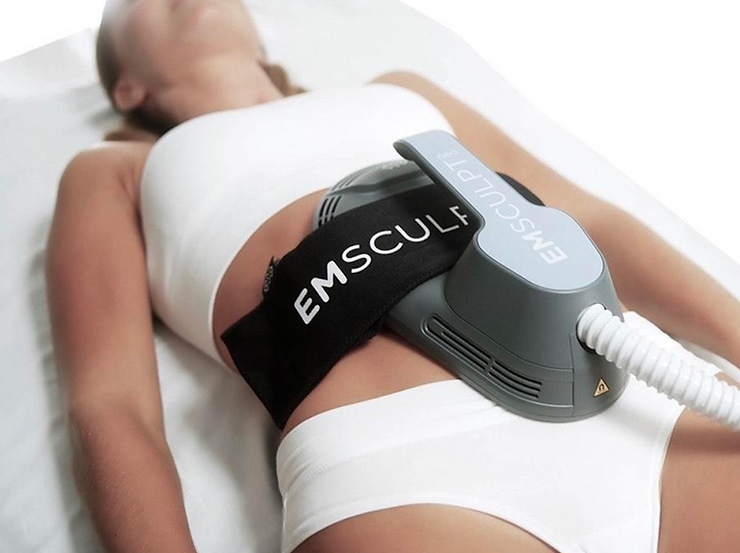 A person is lying down with an EMSculpt device strapped to their abdomen, suggesting a non-invasive body contouring procedure in a clinical setting.