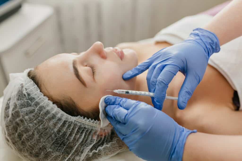 A person is receiving a facial injection from another individual wearing blue gloves in a clinical setting, possibly a cosmetic or medical procedure.