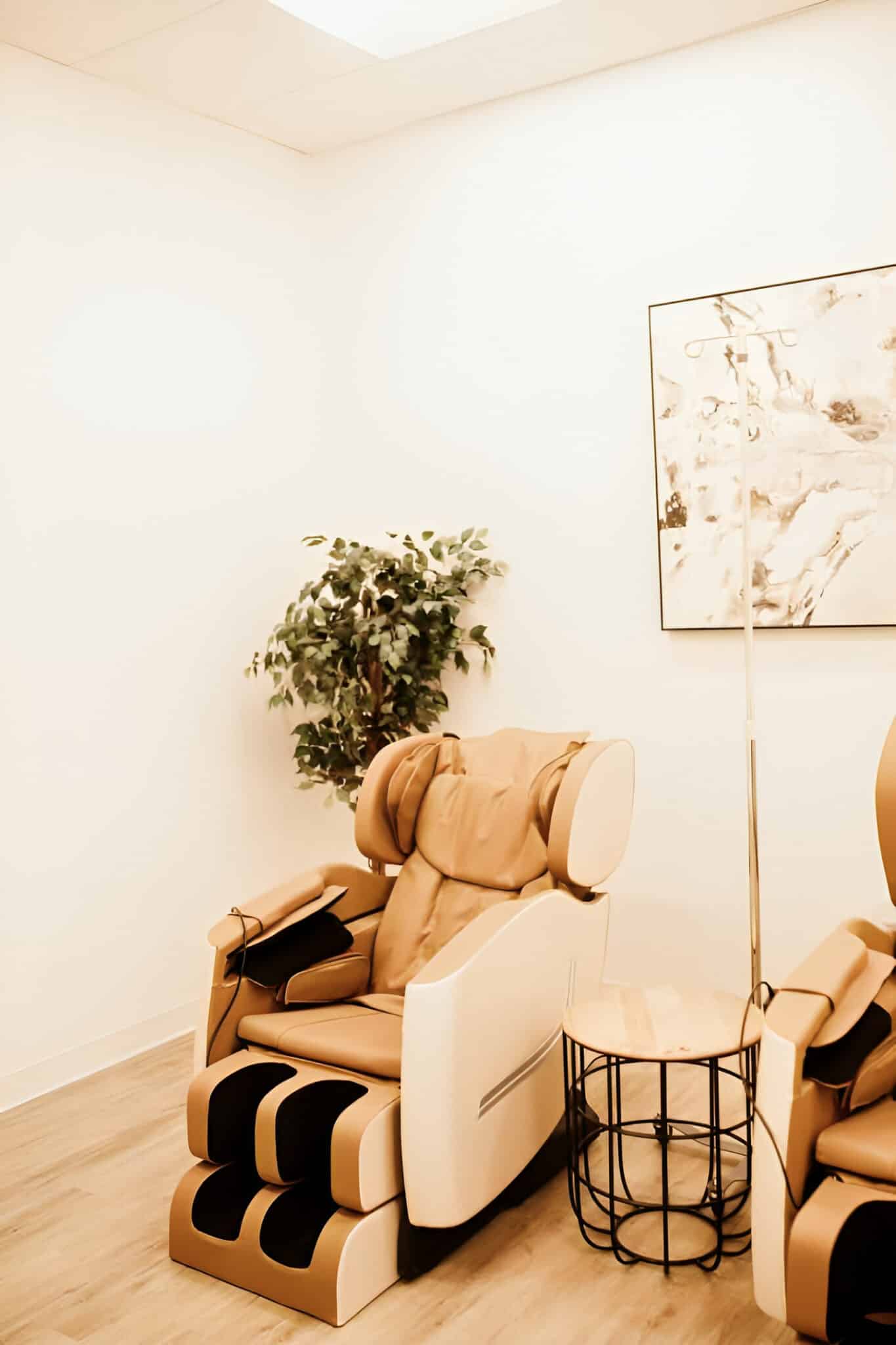 A modern, monochrome room featuring a luxurious massage chair, a small side table, a potted plant, and abstract wall art in warm tones.