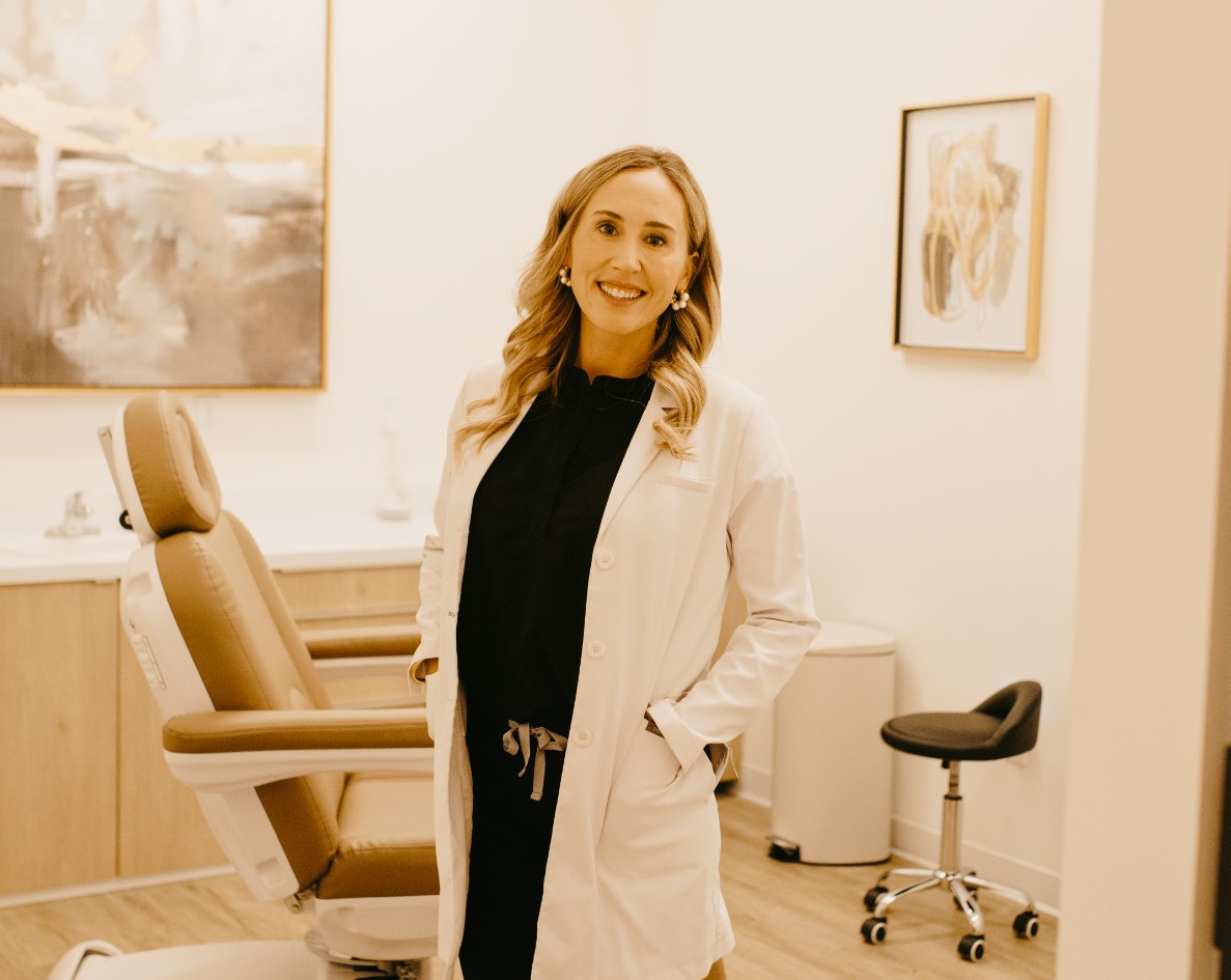 A smiling person in a white coat stands confidently in a dental clinic beside a patient chair, with artwork on the walls in a warm-toned interior.